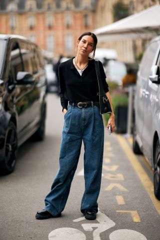 A woman at Fashion Week wearing blue baggy jeans, a black sweater, black shoes, and belt.