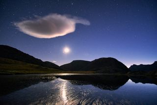 stars in the sky are reflected in the lake below, silhouetted hills are in the background and a large white cloud is present in the sky to the left of the image.