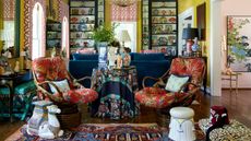 maximalist living room with yellow walls, book cases filled with antiques, large blue couch, red upholstered antique chairs, ceramic stool and leopard bench