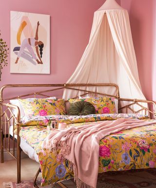 A dusky pink bedroom with a yoga wall art print, a lamp with white drapes, and a rose gold bed with yellow bedding, a pink throw, and green circular throw pillow, with a purple patterned rug underneath