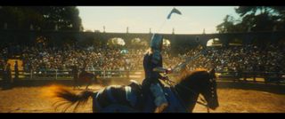 A man in medieval armor rides a horse in front of a crowd in an outdoor stadium, in 'Ren Faire.'