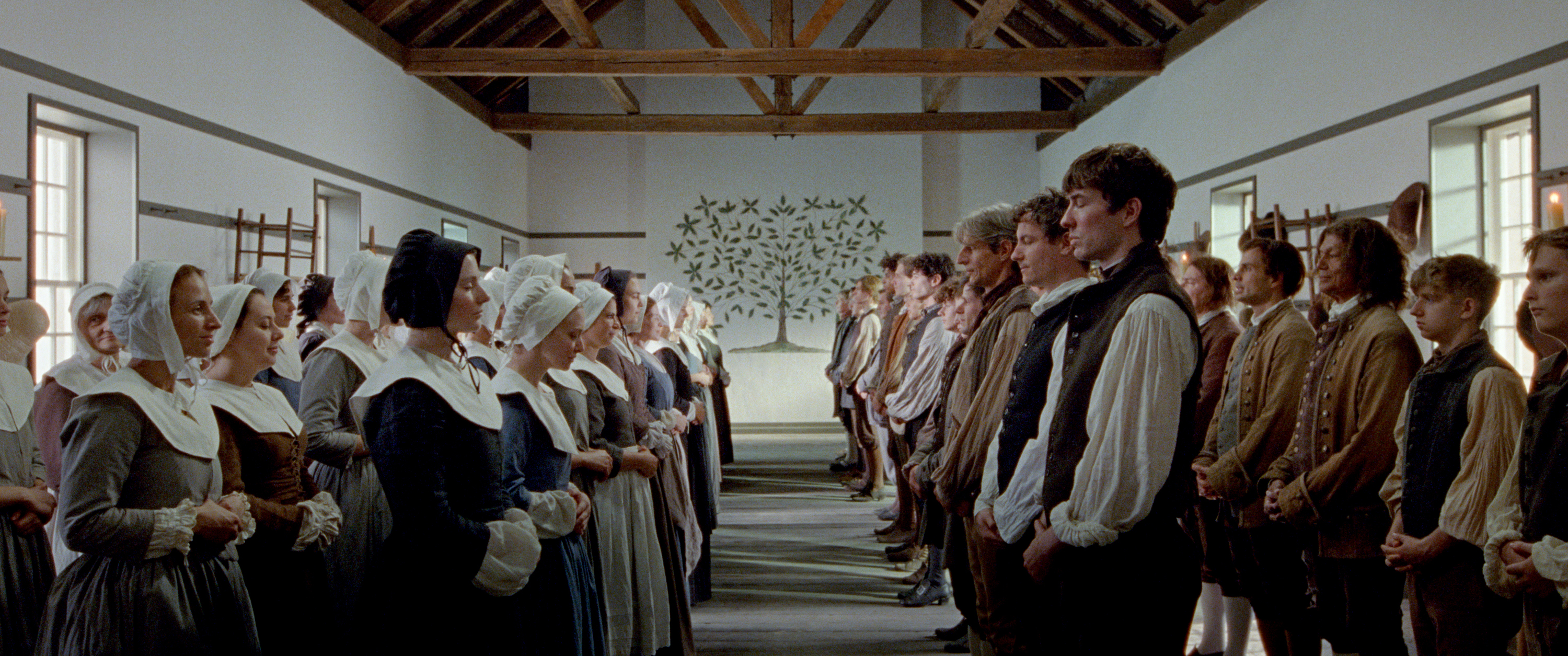 Dozens of Shakers assemble in a meetinghouse, three rows of women in bonnets on the right facing two rows of men on the left. They stand, making a middle aisle leading to a tree painted on the back wall. A still from 'The Testament of Ann Lee.'