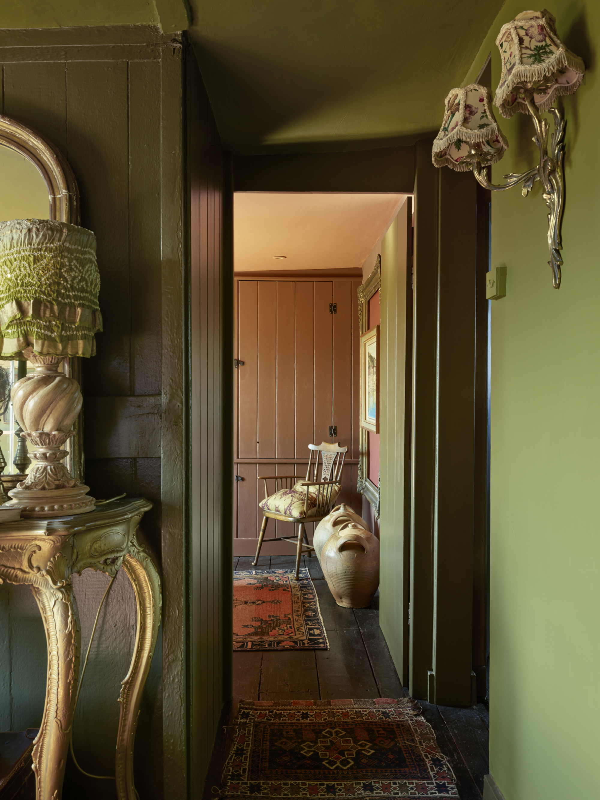 green hallway with vintage rugs, ornate console table and lamps, wall sconces, and pink cupboards in the background with timber chair and vases on the floor