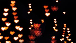 A blurred background filled with glowing heart-shaped bokeh lights in warm hues of orange, red, and white against a dark backdrop