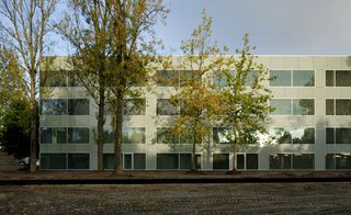 Daytime, exterior image of Campus Hoogvliet, Rotterdam, white flat roof building, multiple windows, tall trees, gravel and soil terrain pathway around the building, blue sky