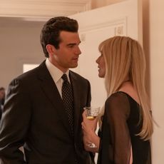 A man in a suit (Paul Anthony Kelly as JFK Jr.) stands with a blonde woman in a black top (Dree Hemingway as Daryl Hannah), who holds a champagne glass. The pair stand next to a flower arrangement in a room of a grand house, in a still from 'FX's Love Story: John F. Kennedy Jr. & Carolyn Bessette.'
