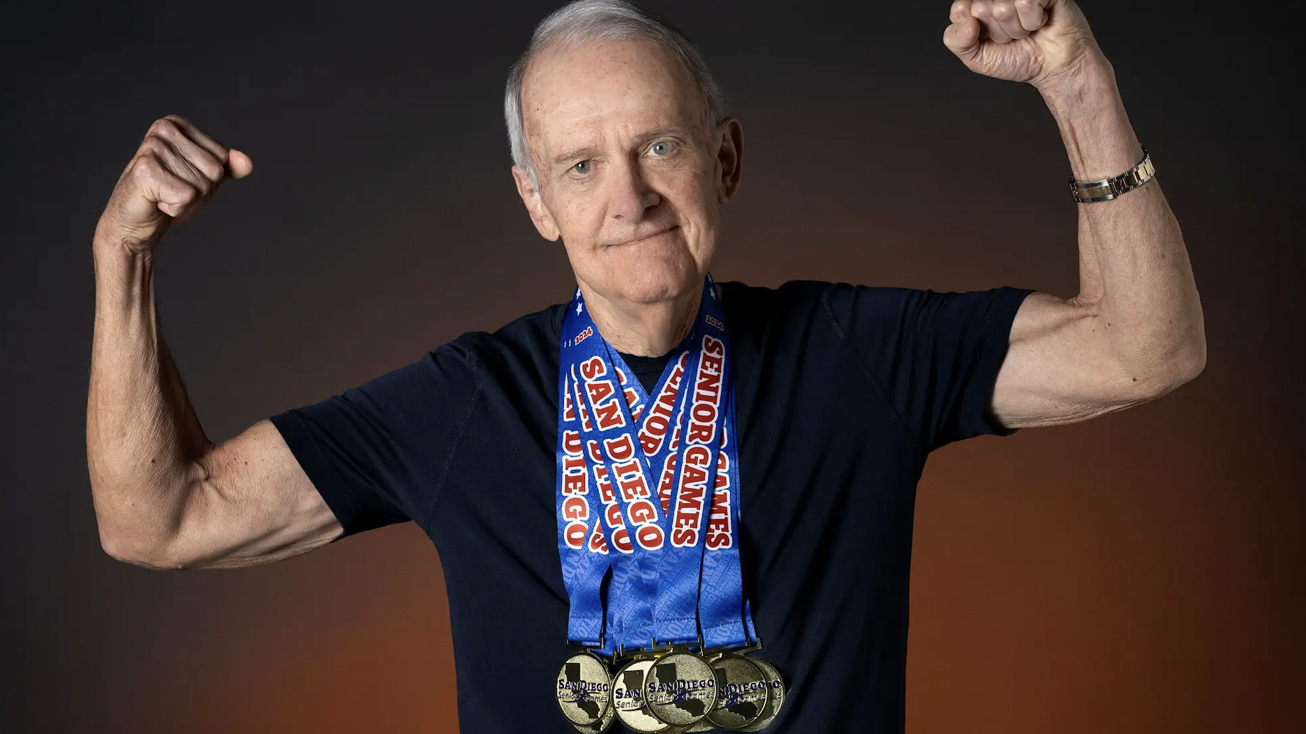 Senior man with arms raised wearing multiple gold medals around his neck, the blue ribbons read &ldquo;San Diego Senior Games&rdquo;
