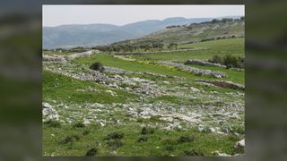 Photograph of Mount Ebal. Gray stone ruins before mountains amid green grass.