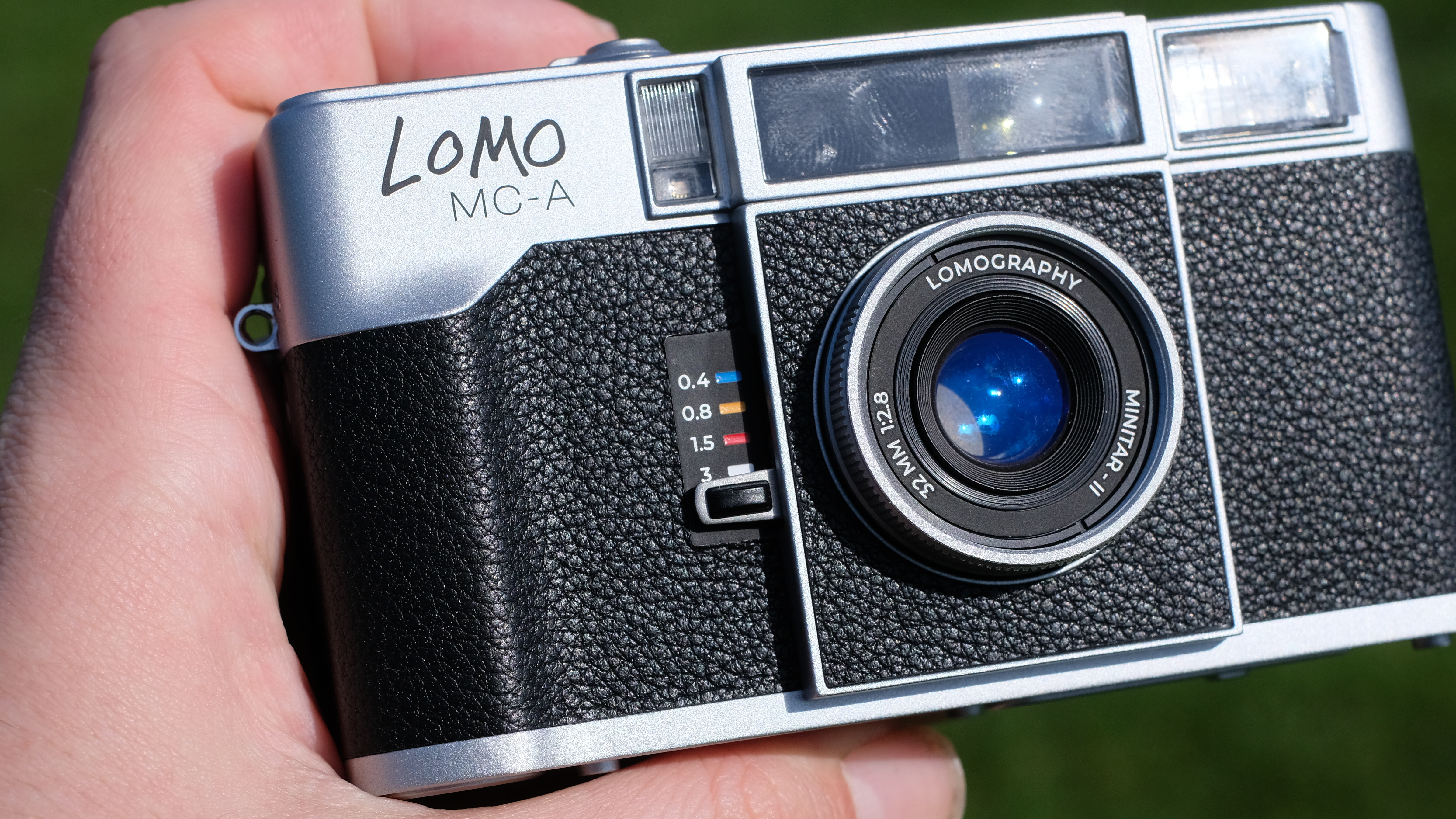 Photograph of the Lomography Lomo MC-A, with grass visible in background, showing switch to set focusing distance