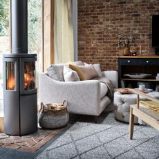 A living room with a wood burning stove and an exposed brick wall and a light grey sofa with a matching ottoman