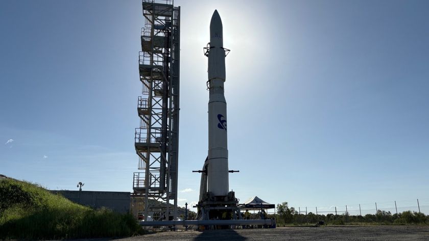 a white rocket with a pointy nose cone stands upright on a launch pad under a bright sun