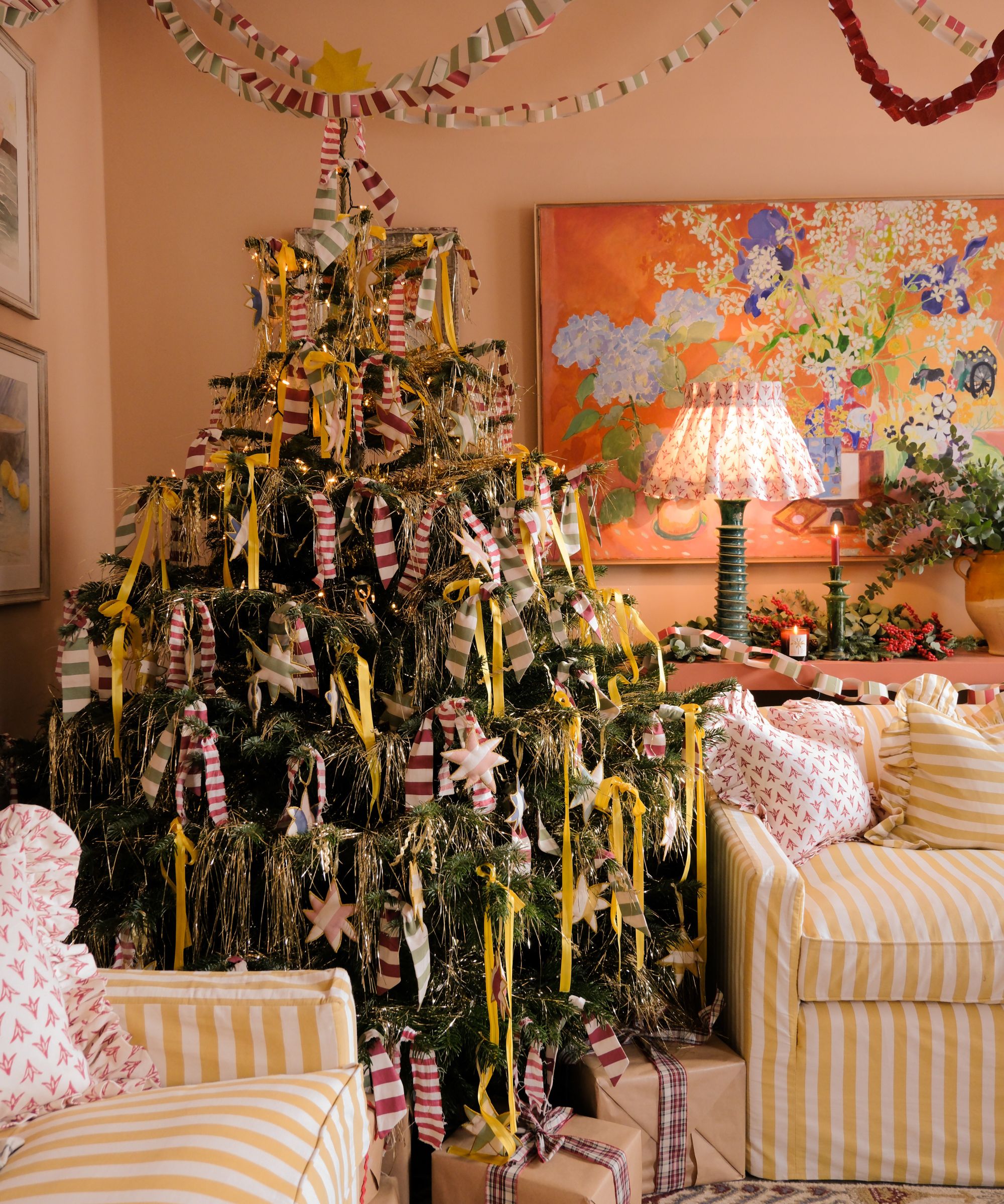 A pale pink living room with yellow striped sofas and a large Christmas tree in the corner decorated with striped bowls, tinsel, and yellow bows. Above are paper chains.