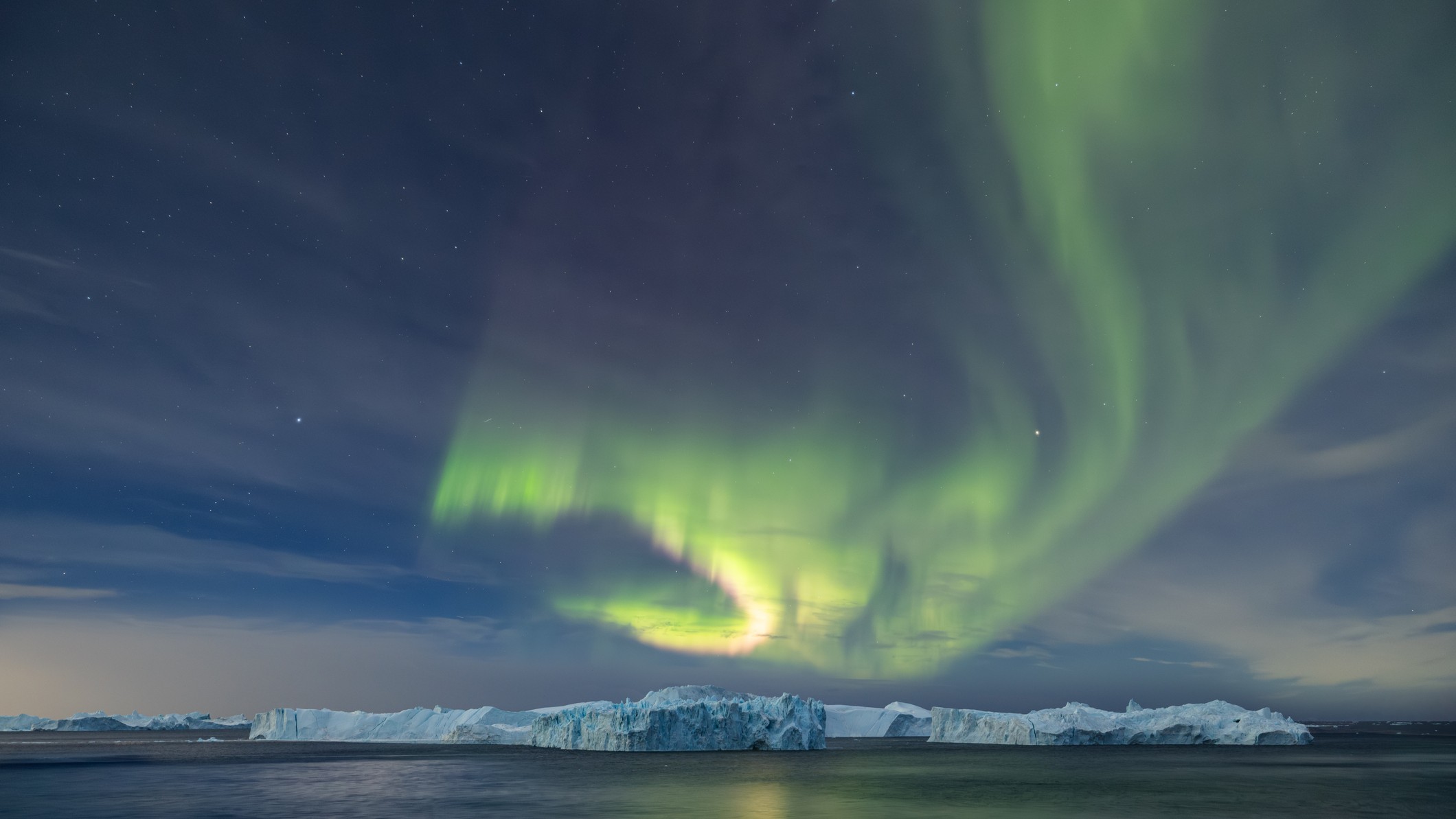 green ribbons of light sweep across the sky above water and a distant ice-covered landmass