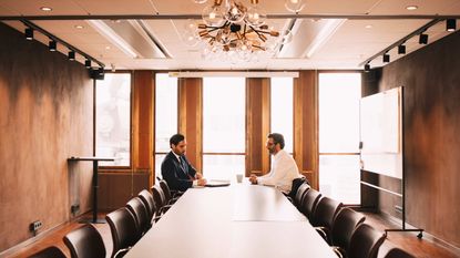 Two men sit across from each other at a long conference room table.