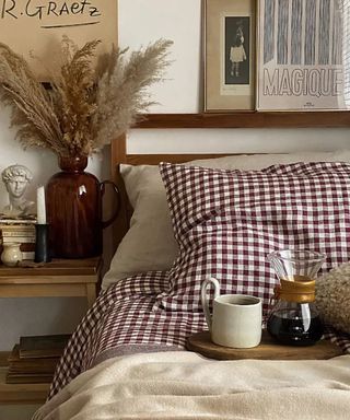 Piglet in Bed Burgundy Gingham sheets on a bed with a breakfast tray; to the left, a white oak nightstand with a vase filled with stems; beige posters against a white wall.