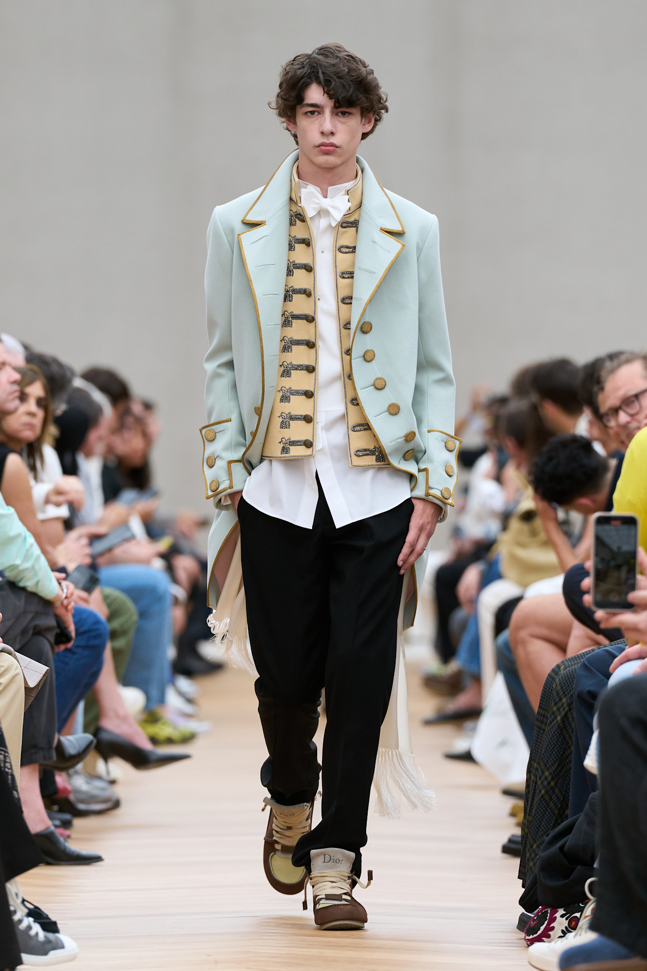 a model at Dior Menswear Spring 2026 wearing a blue frock coat, tan military jacket, white shirt, and dark black pants.