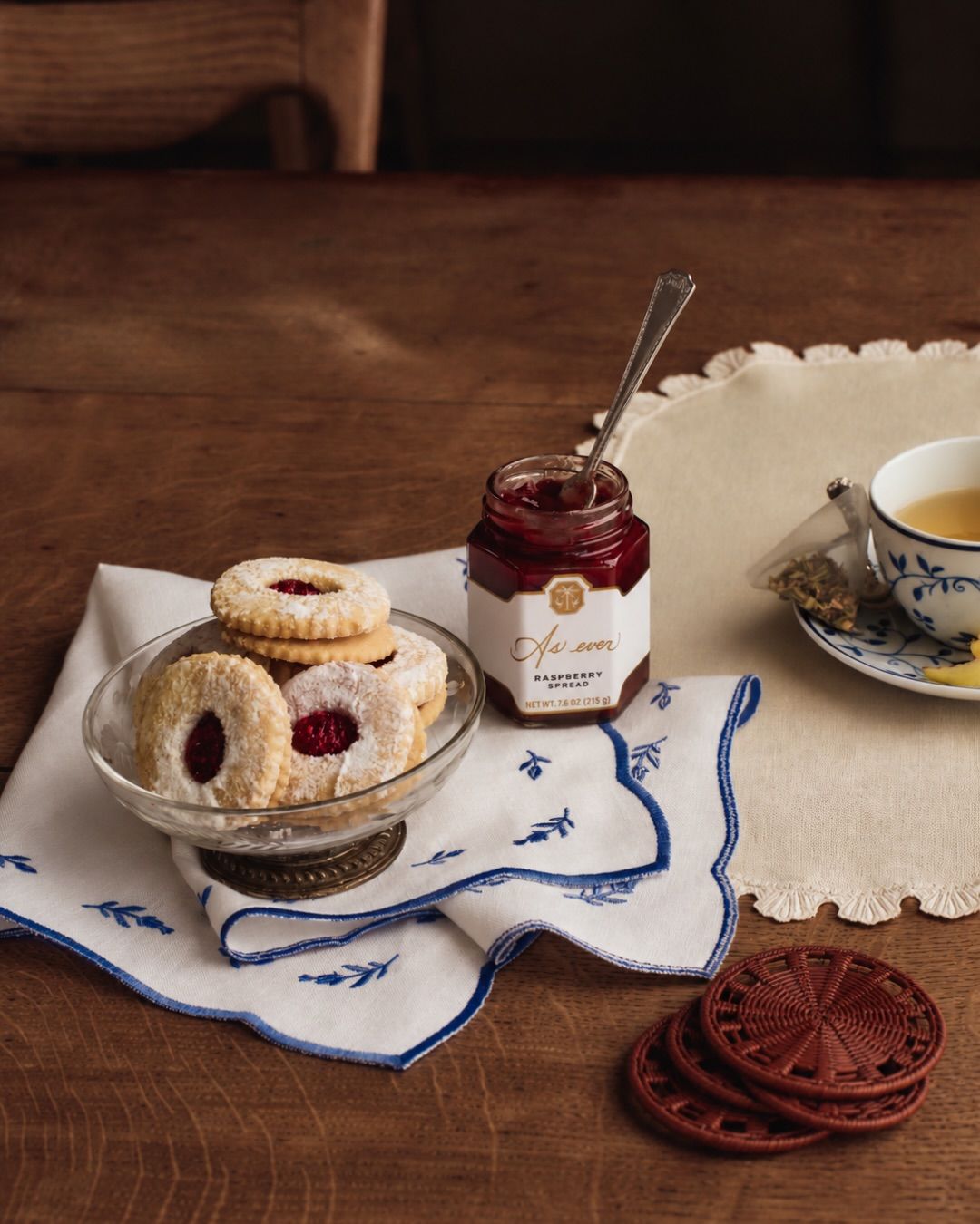 A table with napkins, a bowl of cookies, a white tea cup and raspberry spread