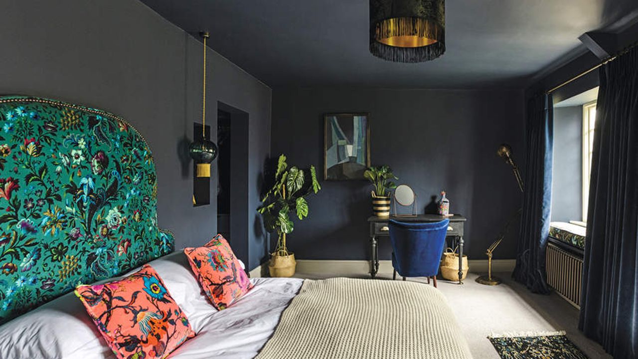 Eclectic and dramatic bedroom with dark blue walls and ceiling, wallpapered feature wall and double bed with white bedding and bright cushions. feature headboard with dramatic and colourful fabric