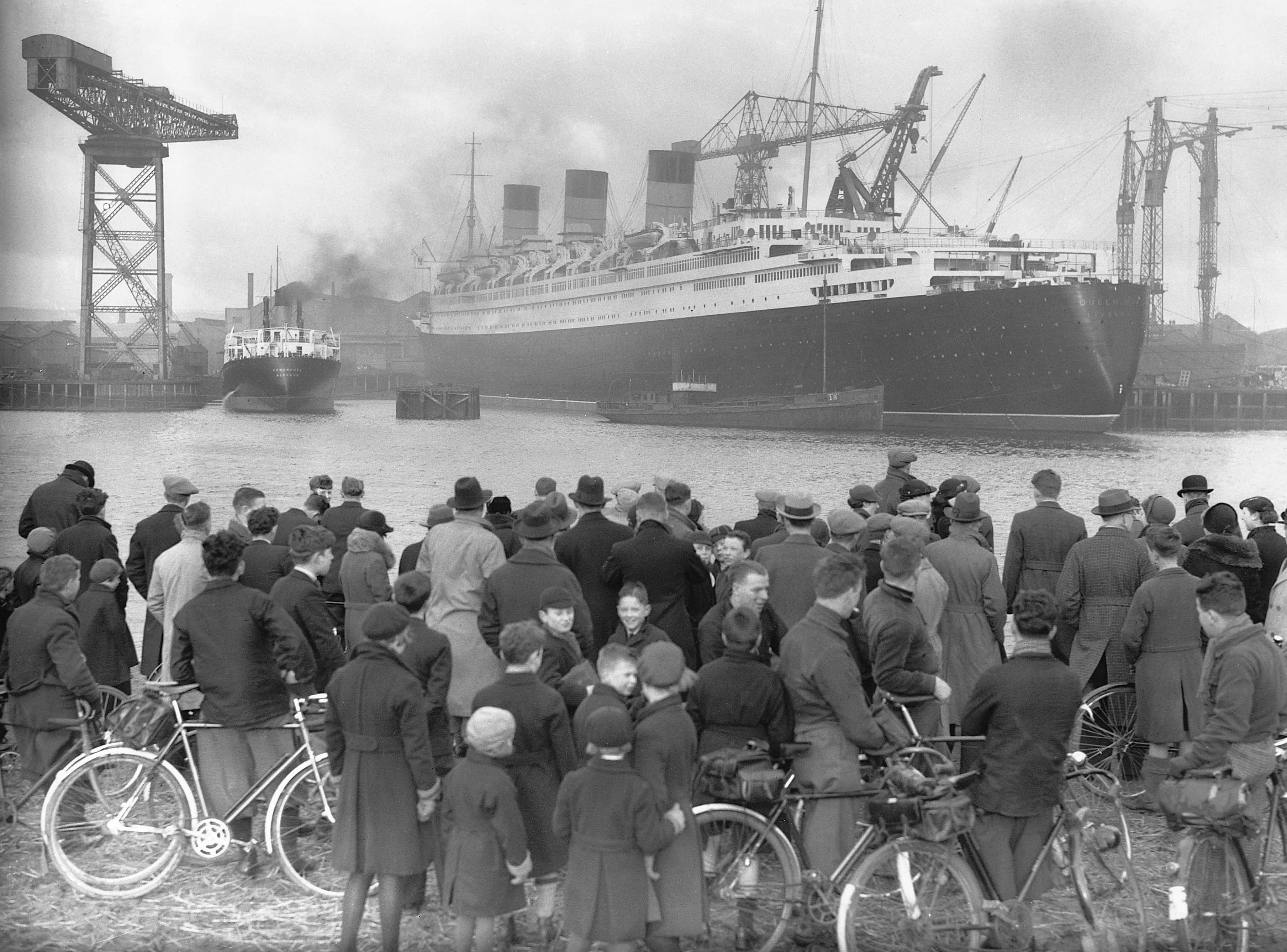 A crowd of men and boys, some with bicycles, stands on a riverbank looking across the water at the huge liner Queen Mary, with a giant crane on the left and a smoking tug alongside.