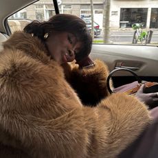 Amy is in a car wearing a fur coat, black bag, and gold earrings.