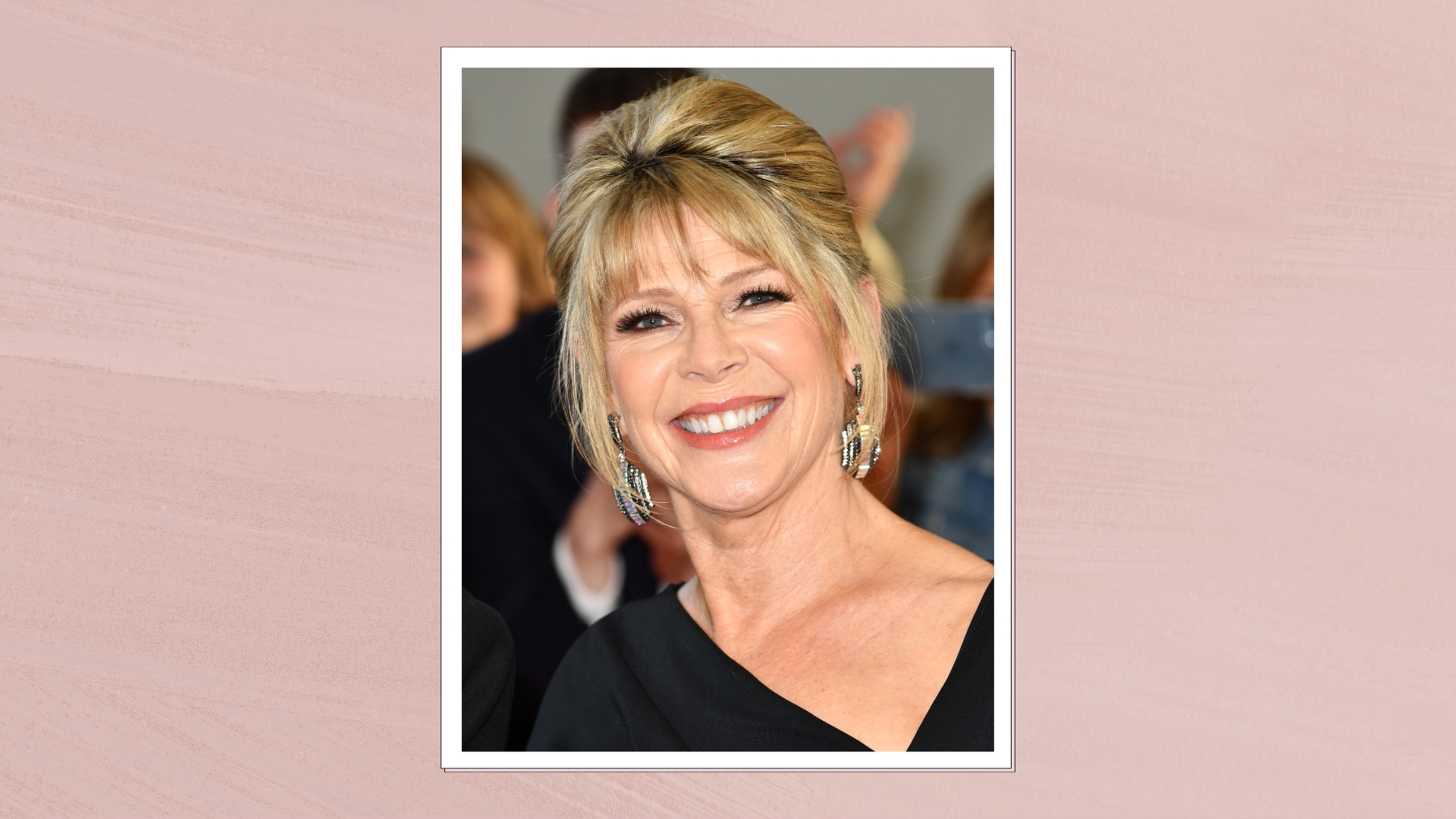 Image of Ruth Langsford smiling with blonde updo and feathered fringe, wearing black gown with asymmetric neckline, on a pink background