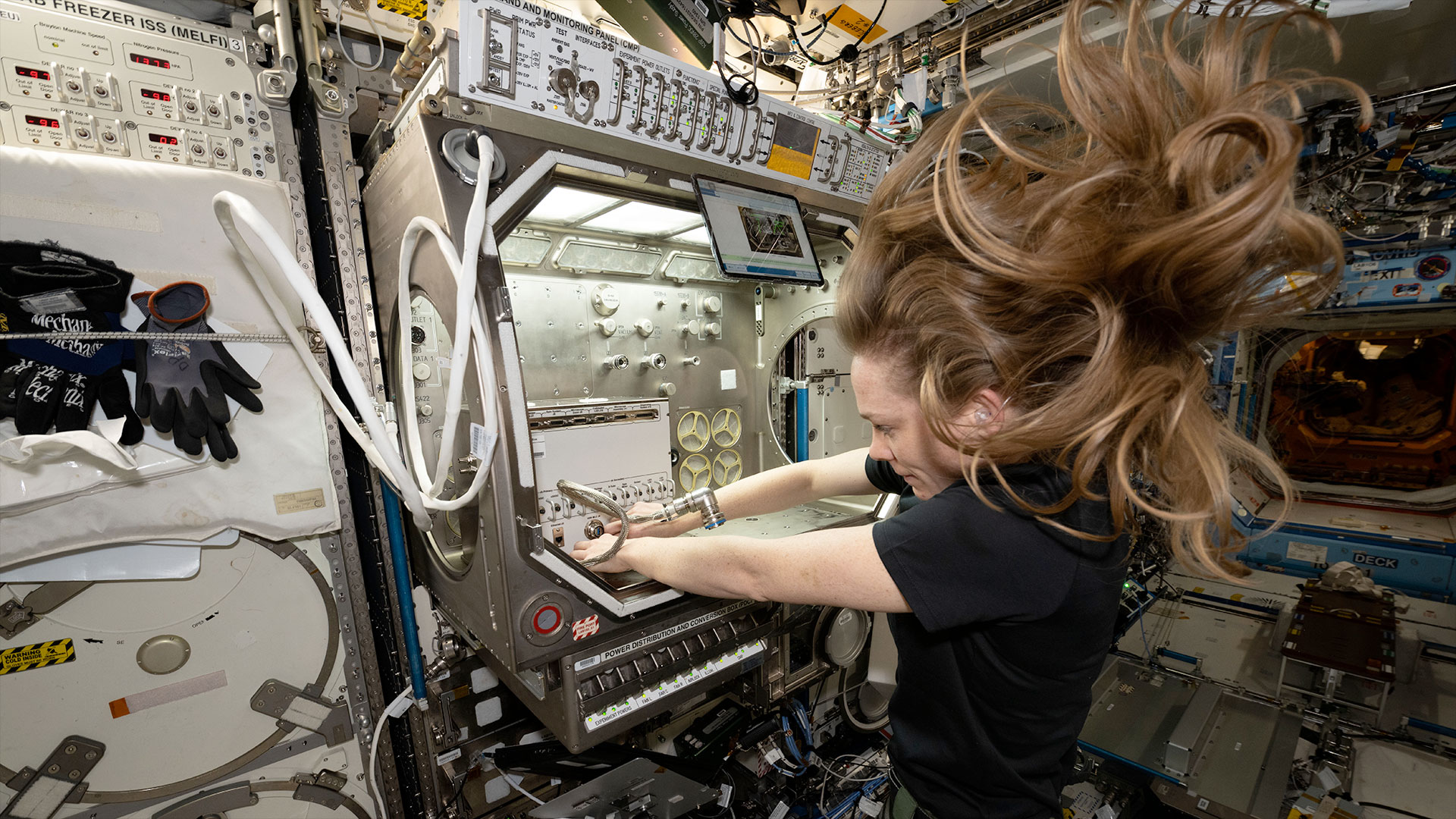 a woman with long brown hair floating freely in microgravity works to install an experiment a science glovebox aboard a space station.