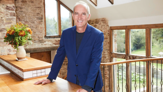 Kevin McCloud, wearing a royal blue jacket, smiles in an airy but rustic cottage kitchen as he presents &quot;Grand Designs: 25 Years and Counting&quot; 