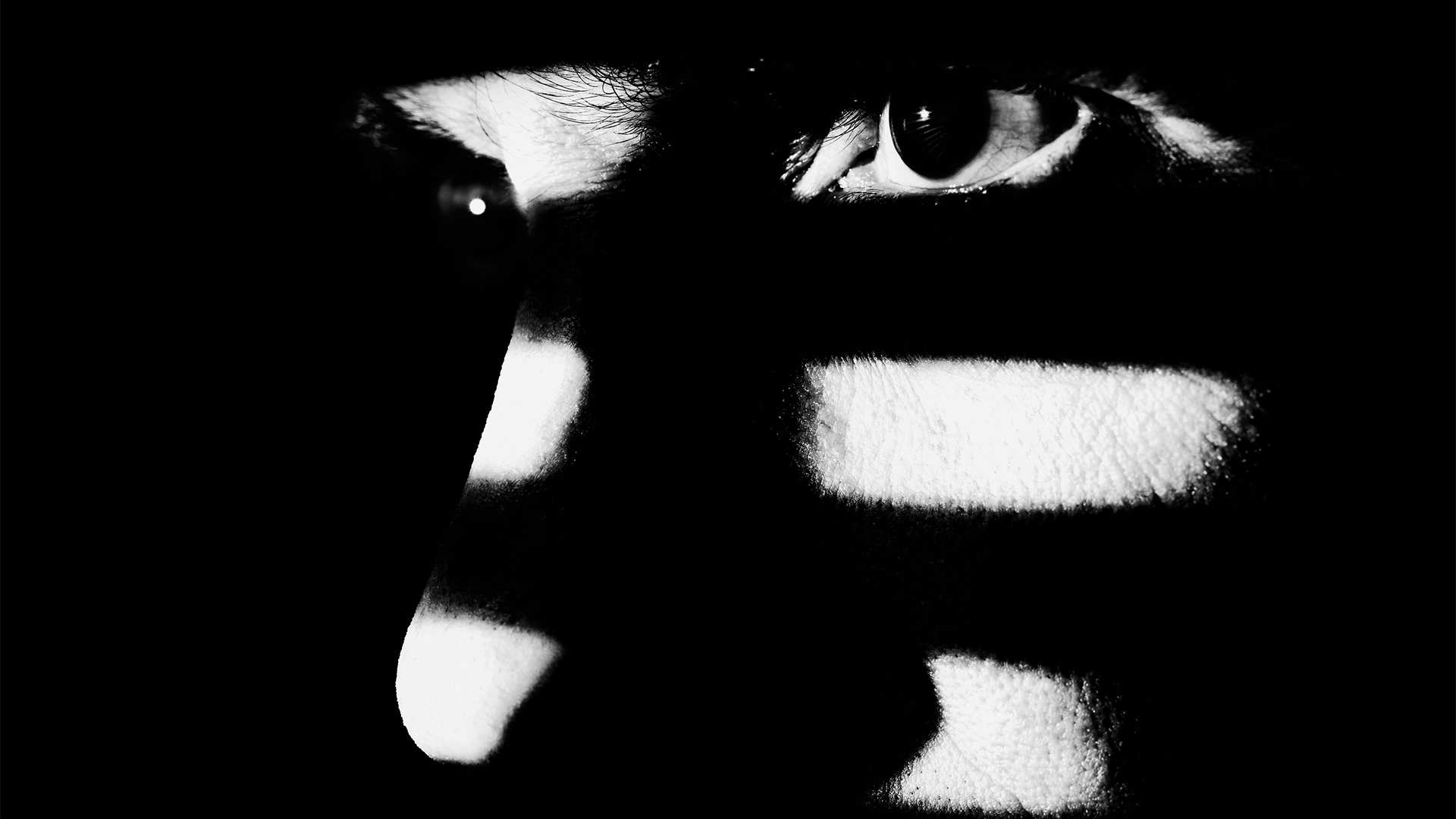 Concept image showing man lurking in shadows with strands of light hitting different parts of his face while staring intently.