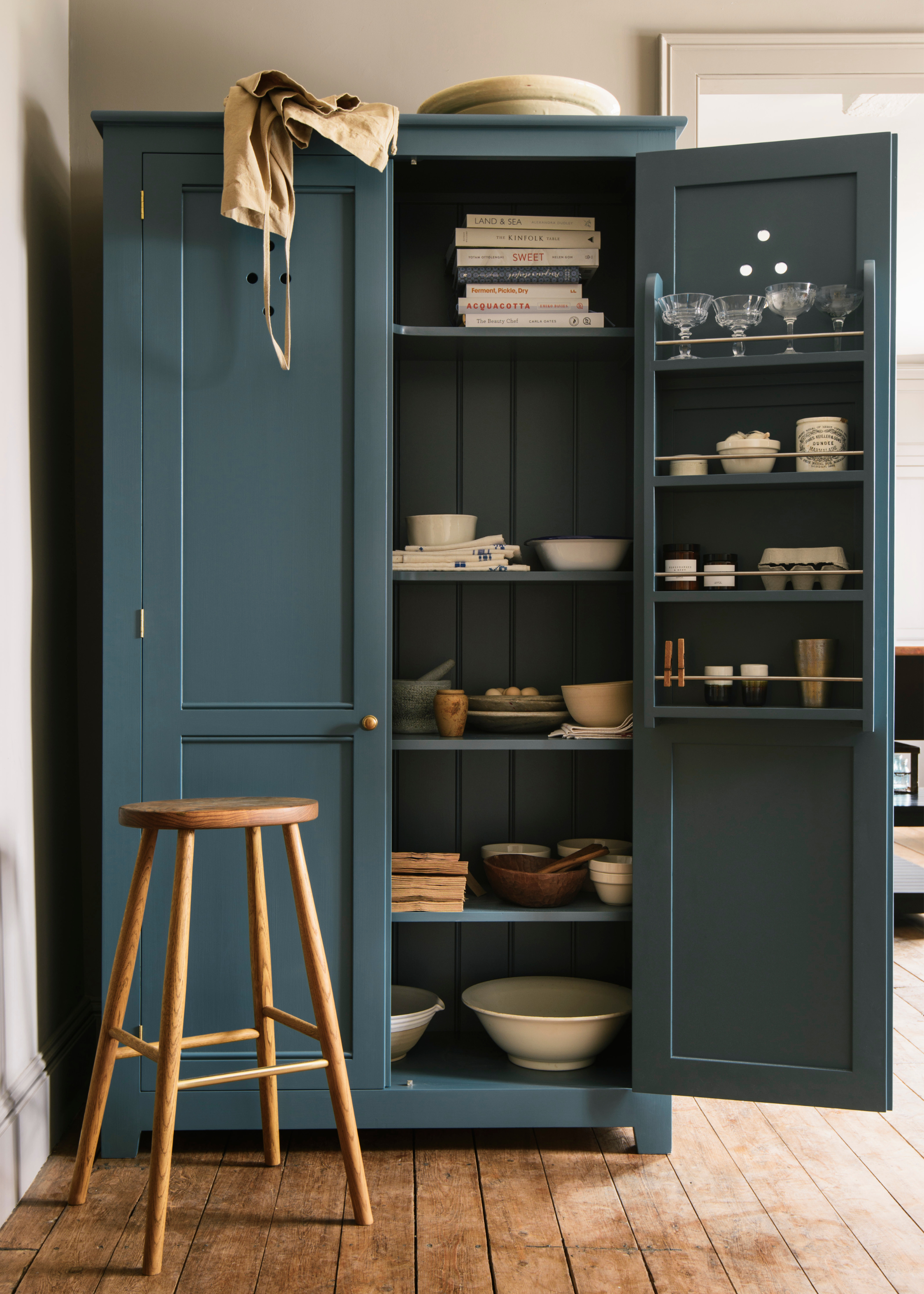 a freestanding pantry in teal with the door open showing internal and door-mounted shelving for dinnerware and glassware with a wooden stool in front