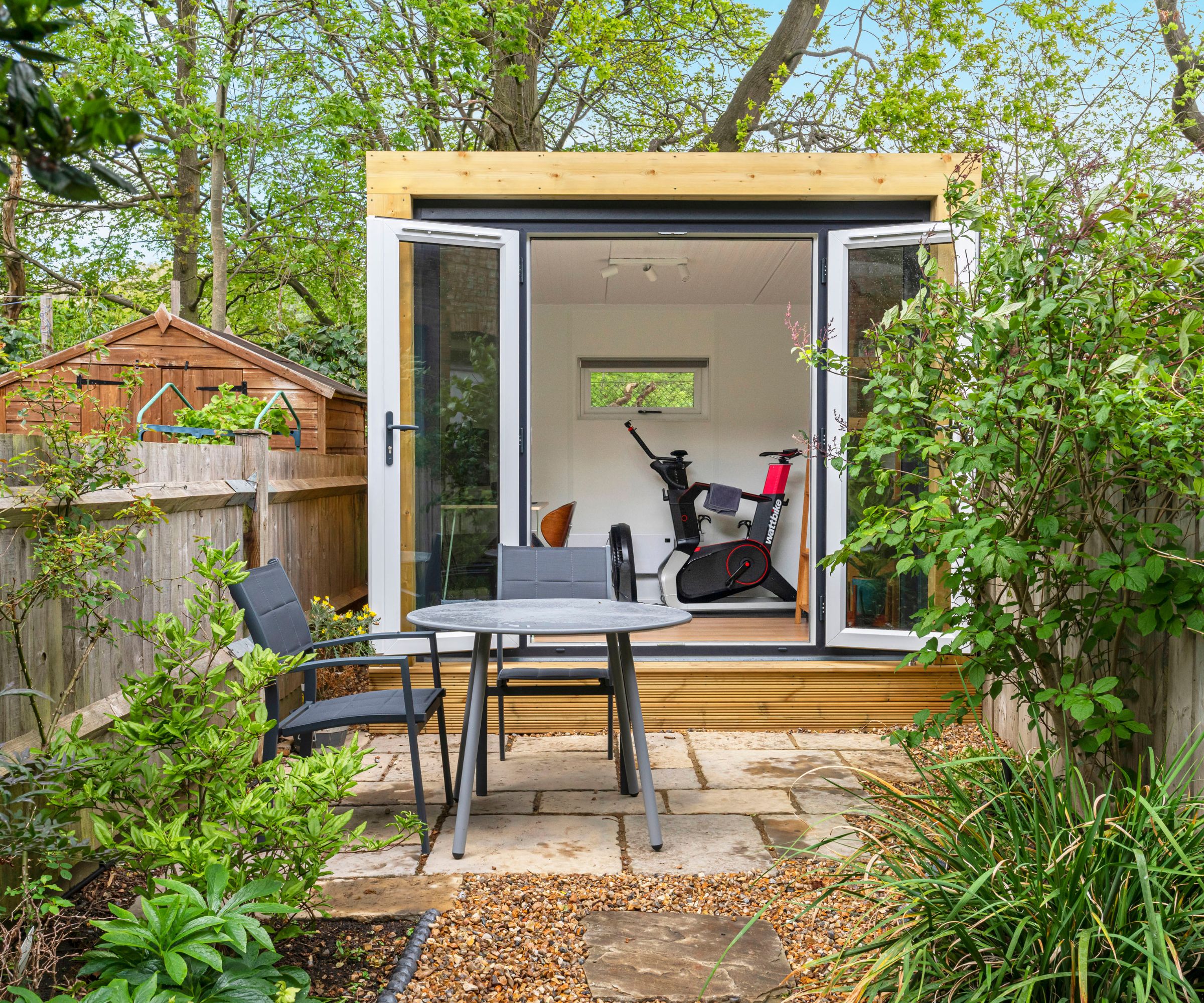 Small timber garden gym with exercise bike visible through open doors, patio table and chairs outside.