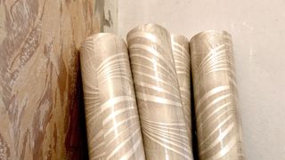 Four rolls of gold patterned wallpaper leant against a wall with brown patterned wallpaper