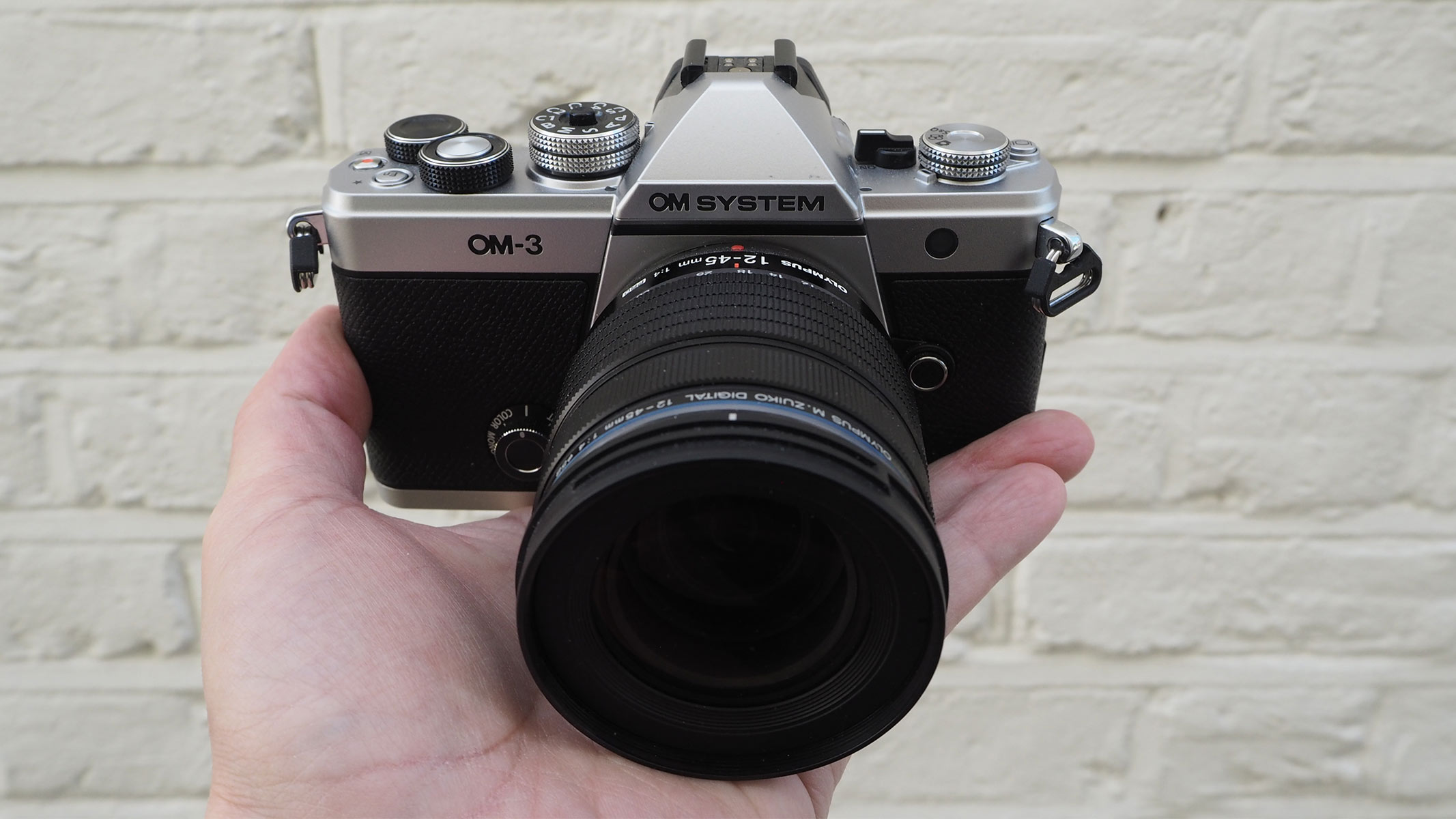 Reviewer holding the OM System OM-3 in his hand against a white brick wall.