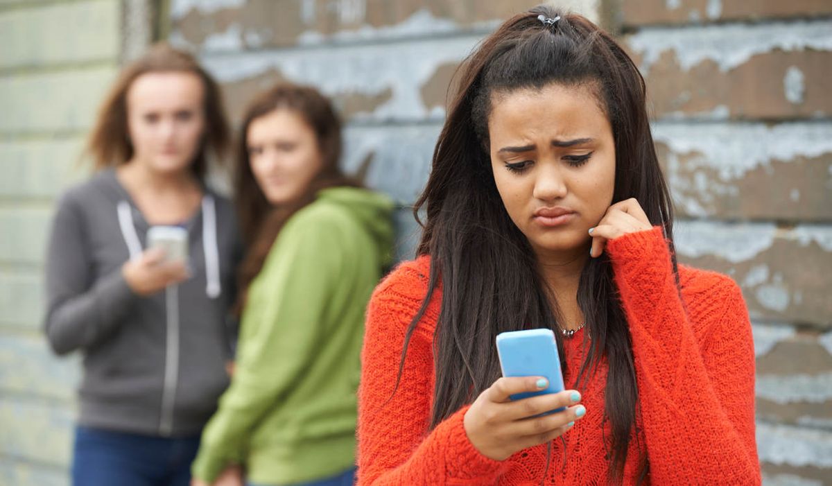 Sad teen girl looks at her cell phone while two girls in the background smirk