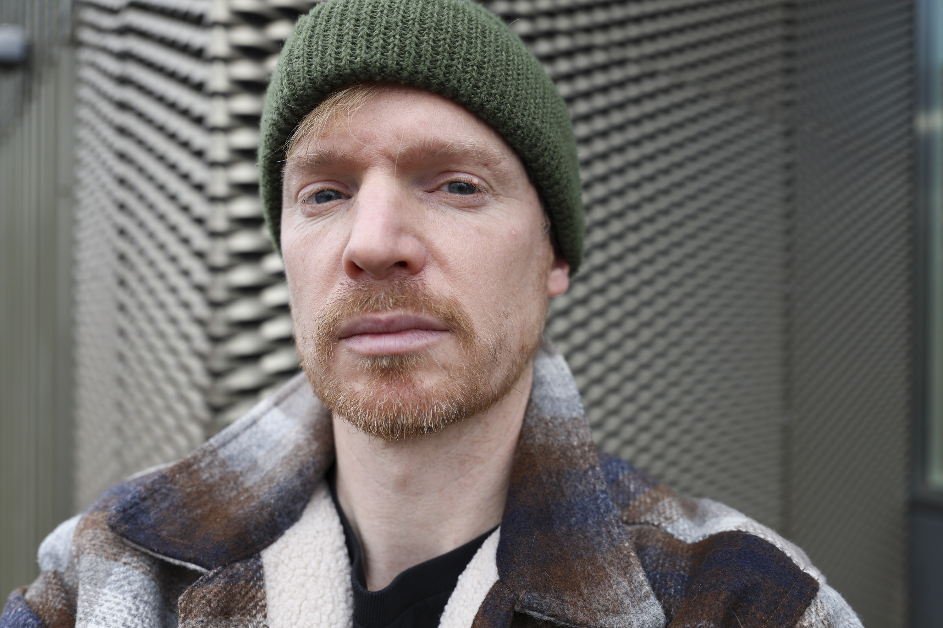 A man in a green beanie and shacket's selfie, outdoors in gray urban area