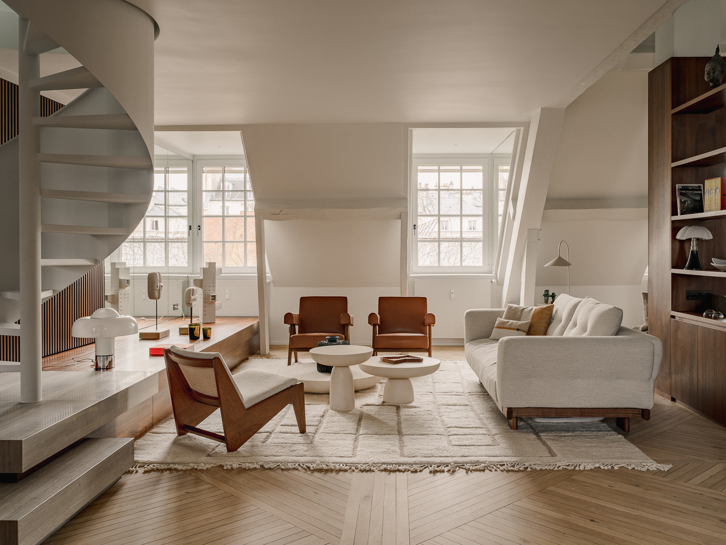 A refined attic apartment interior characterized by a warm, modernist aesthetic. The space is organized as an open-plan living area under a pitched roof, with exposed white-painted beams and triangular dormer windows that bring in soft natural light.