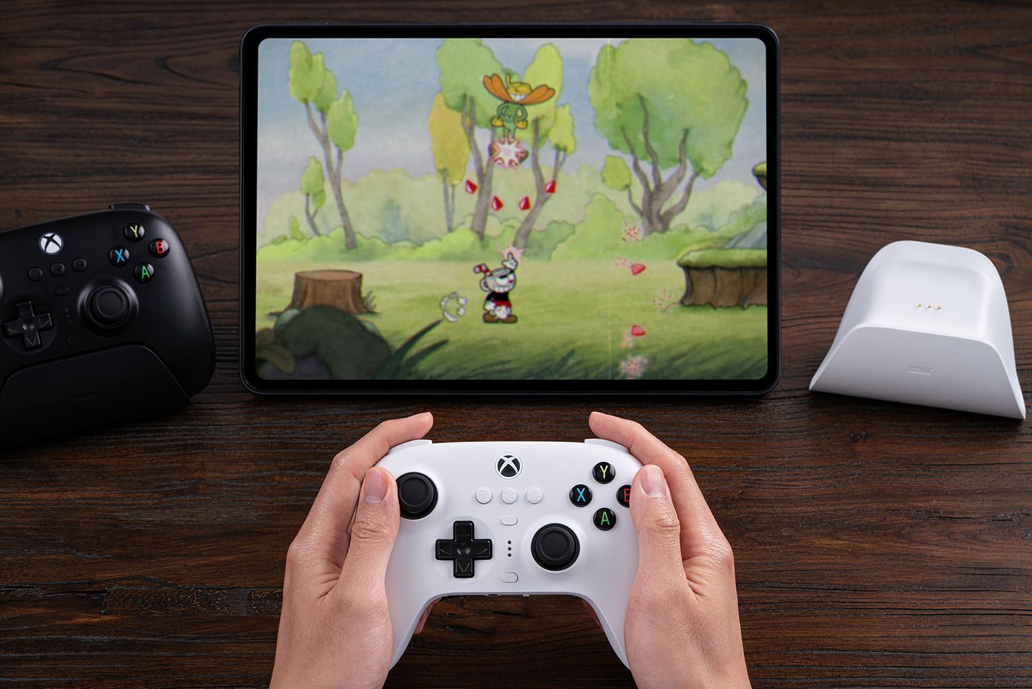 Hands holding a white Xbox controller in front of a tablet displaying a colorful cartoon game with trees. A black controller and charging dock are nearby.