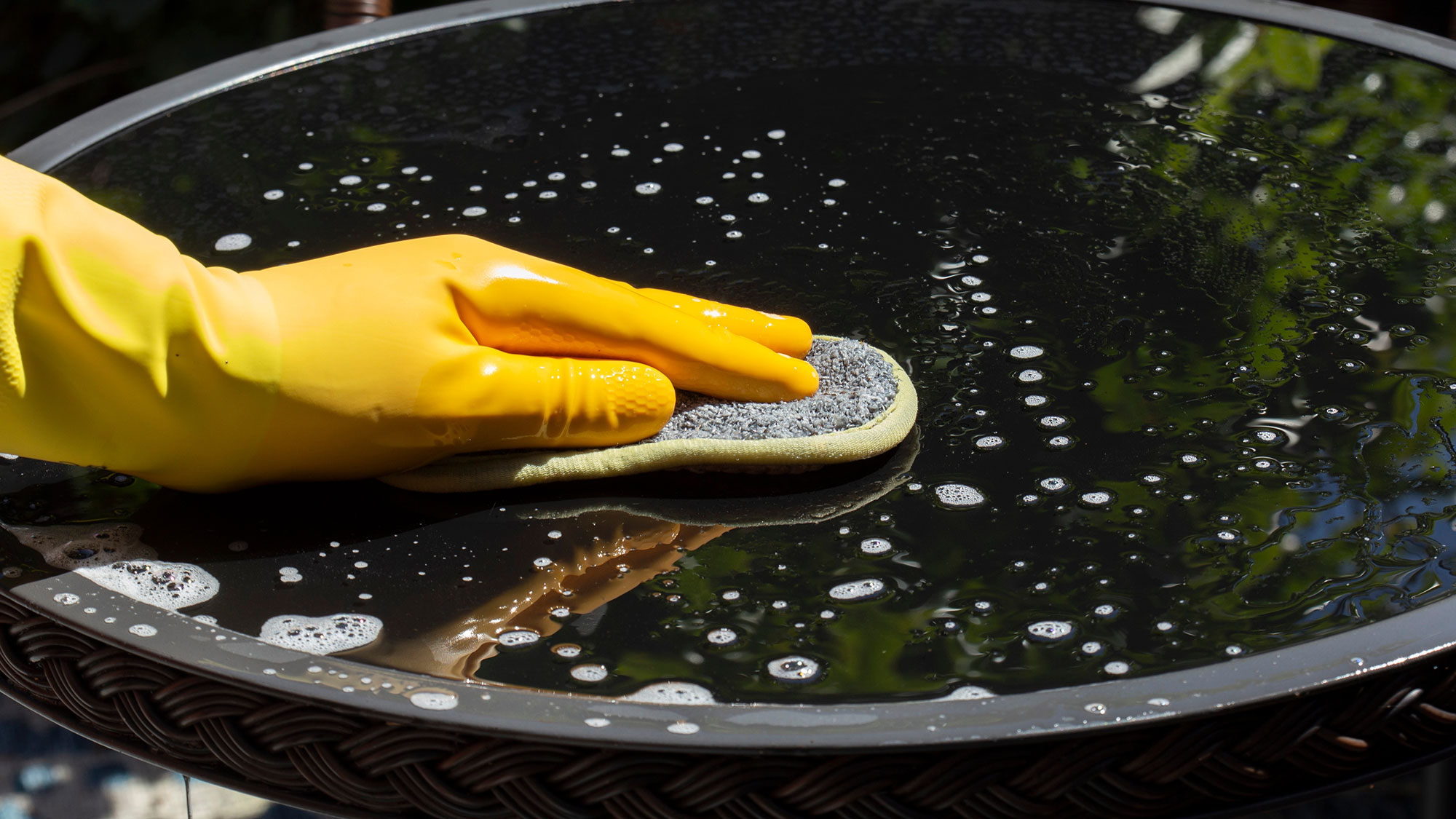 Person wearing rubber gloves cleaning a glass top patio table in a garden backyard