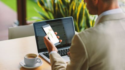 A man looks at a stock trading graph on his phone while sitting at a cafe table with his laptop open.