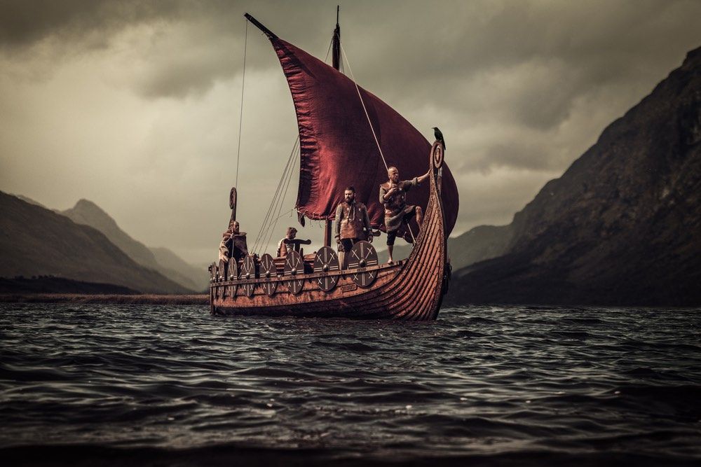 Group of vikings are floating on the sea on Drakkar with mountains in the background.