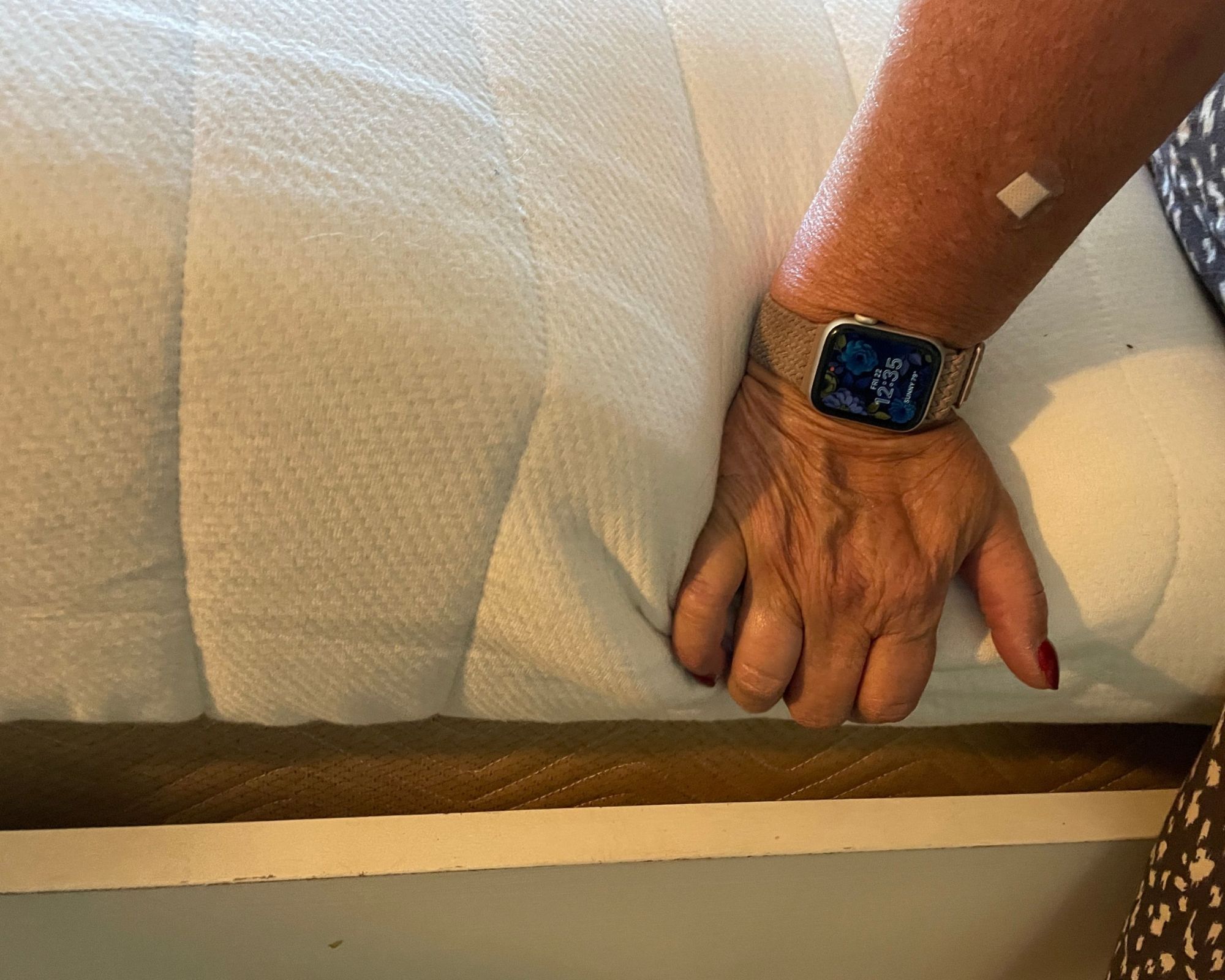 An arm and hand testing the edge support on the white Earthfoam Organic Mattress. On the arm is a small circular clear plaster, and on the wrist is an Apple watch with a floral background and the time '12:35' displayed on a beige strap.