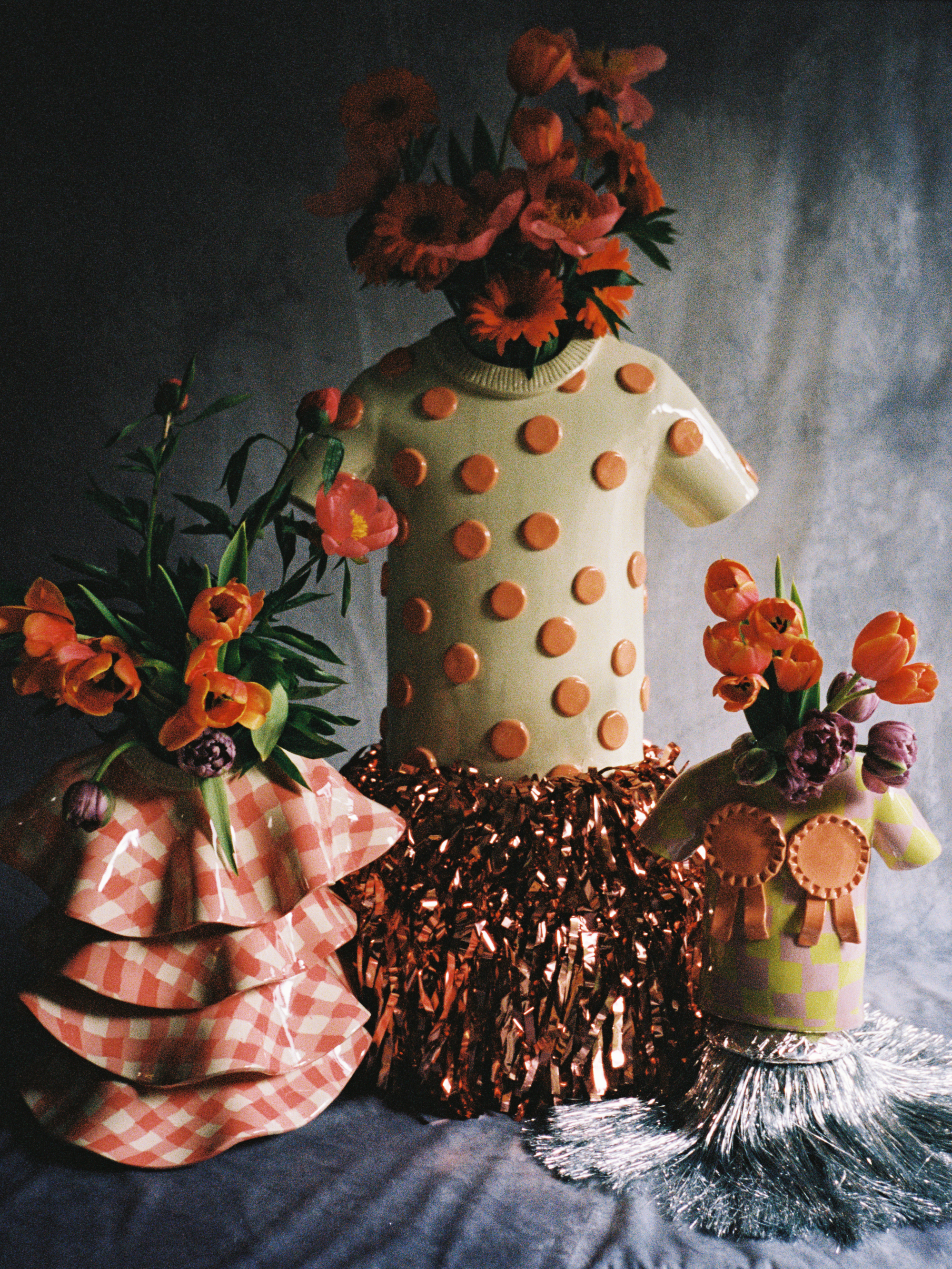 Image of three vases that are shaped like colorful t-shirts. there are flowers in each of the vases and the background is a gray tie-dye color.