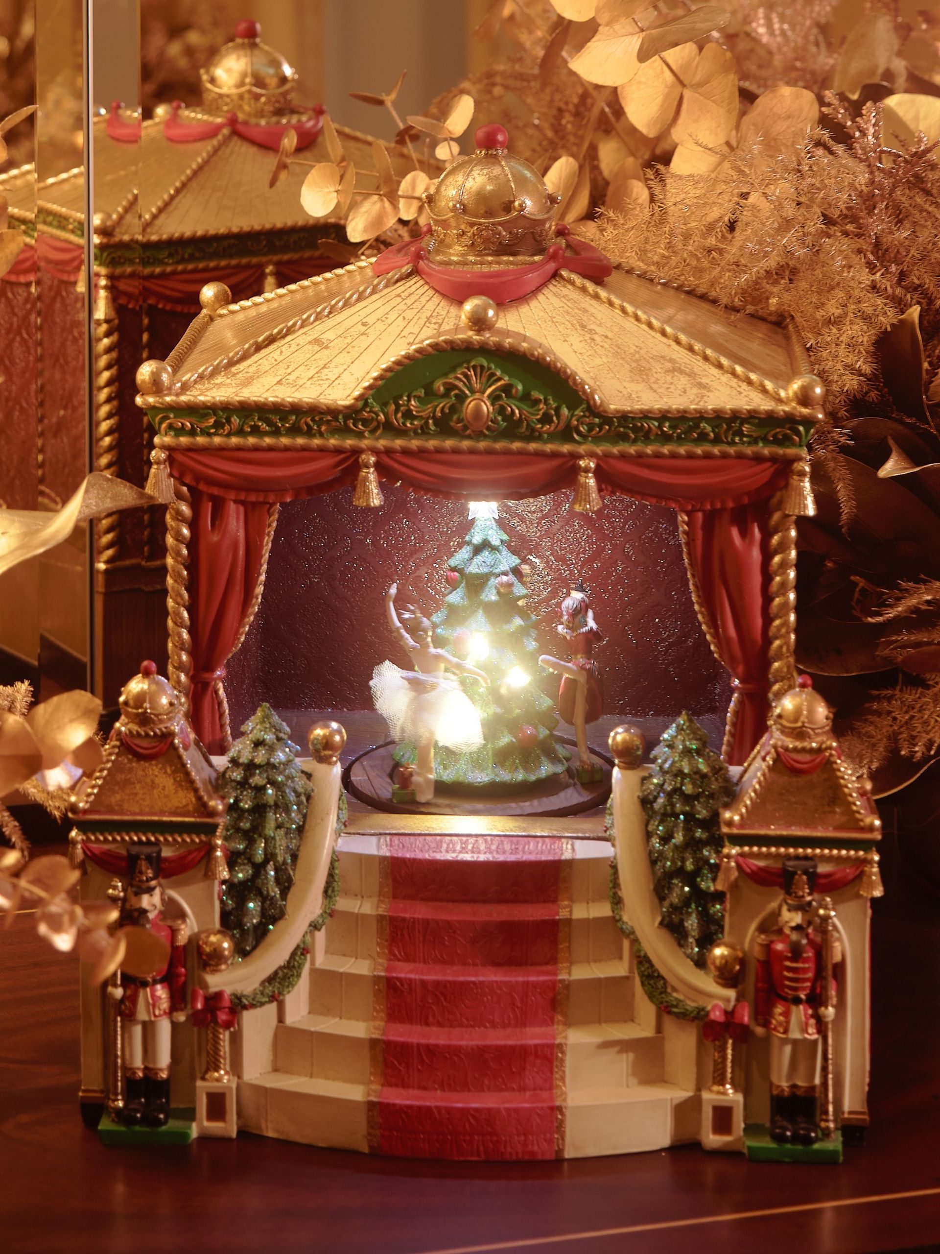 A miniature theatre with dancers on the stage dancing around a Christmas tree on a mantel in among gold dried floral arrangements and lights