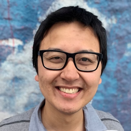 Image of a man in black glasses and a gray collared shirt standing in front of a blue background.