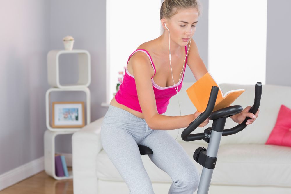 A woman reads a book while riding a stationary bike.