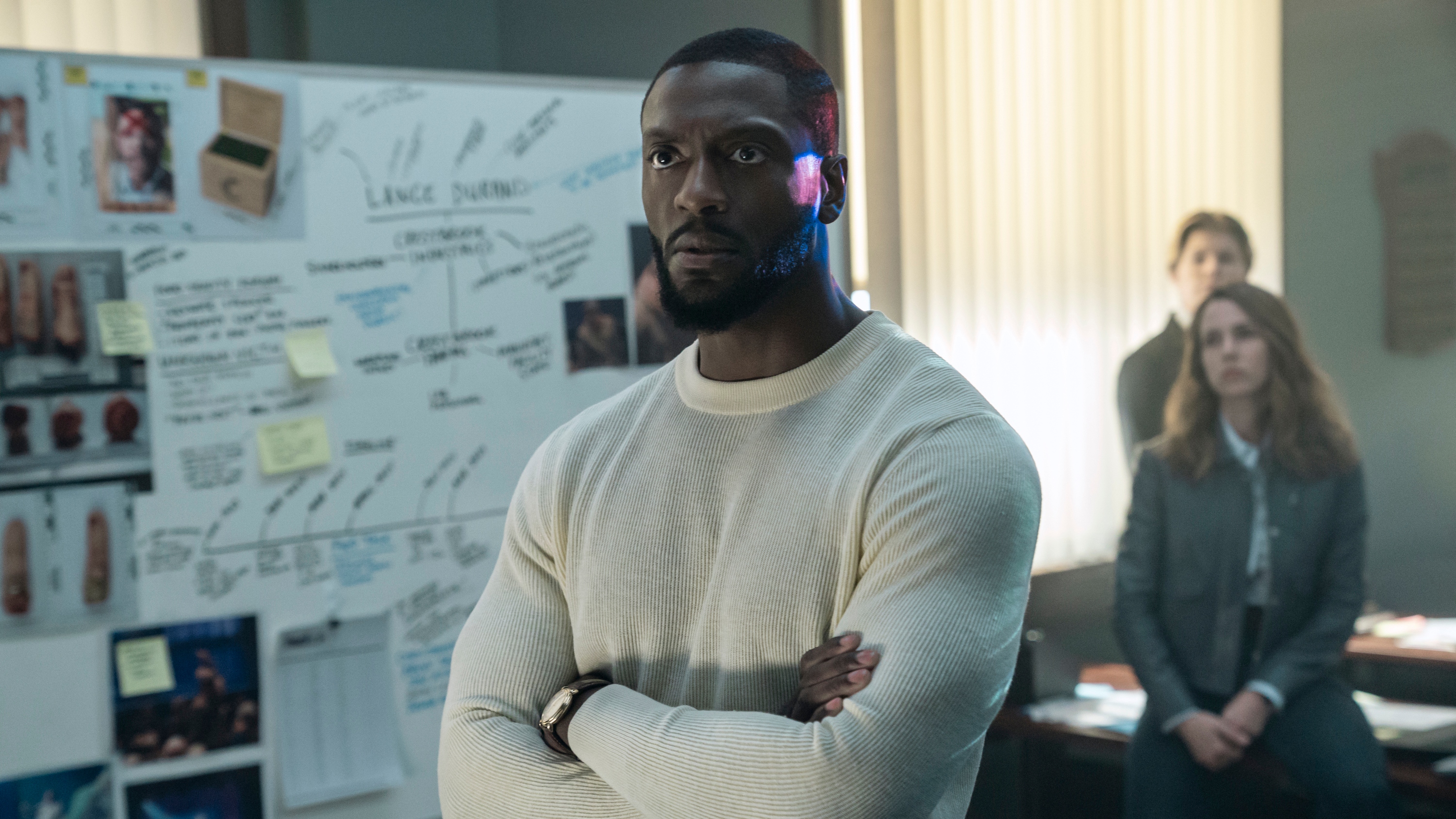 Aldis Hodge as Alex Cross in a white jumper with his arms folded, standing in front of a whiteboard with notes about a murder on it, while colleagues look on