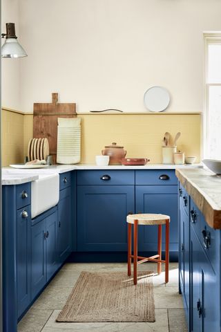 A kitchen with blue cabinetry furniture and yellow walls and wooden stool seating, stone flooring and jute rug by Little Greene