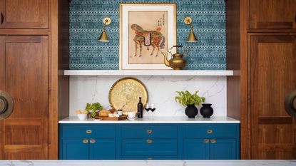 Lighting trends are so chic. Here is a kitchen with a blue wallpapered wall with two gold wall sconces, dark blue cabinets with white countertops above with a cheese board and two black vases, and a marble island