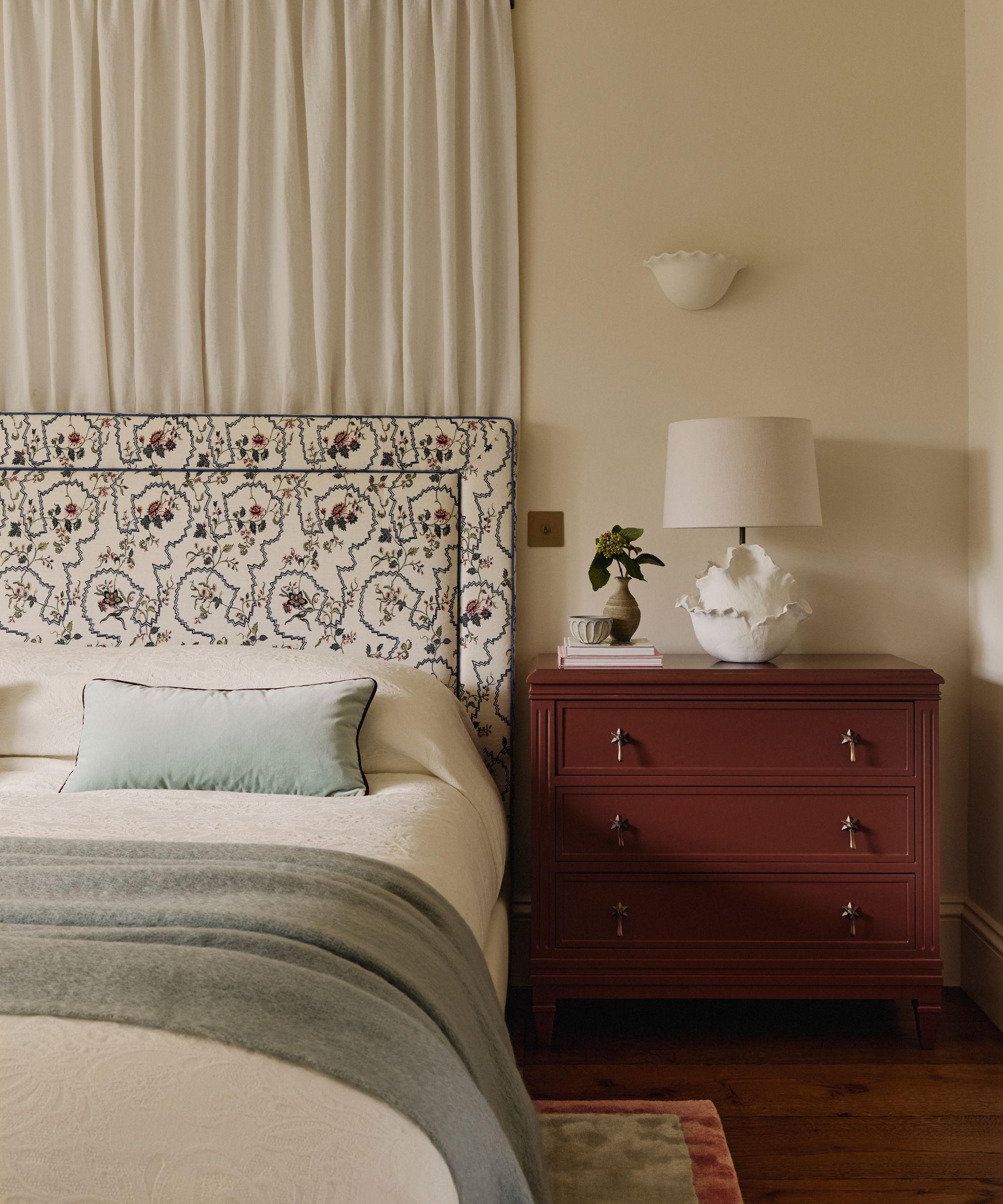 neutral bedroom with a curtain hung behind the bed, a floral headboard and neutral linens with a dark red painted nightstand styled with a lamp, vase, and books