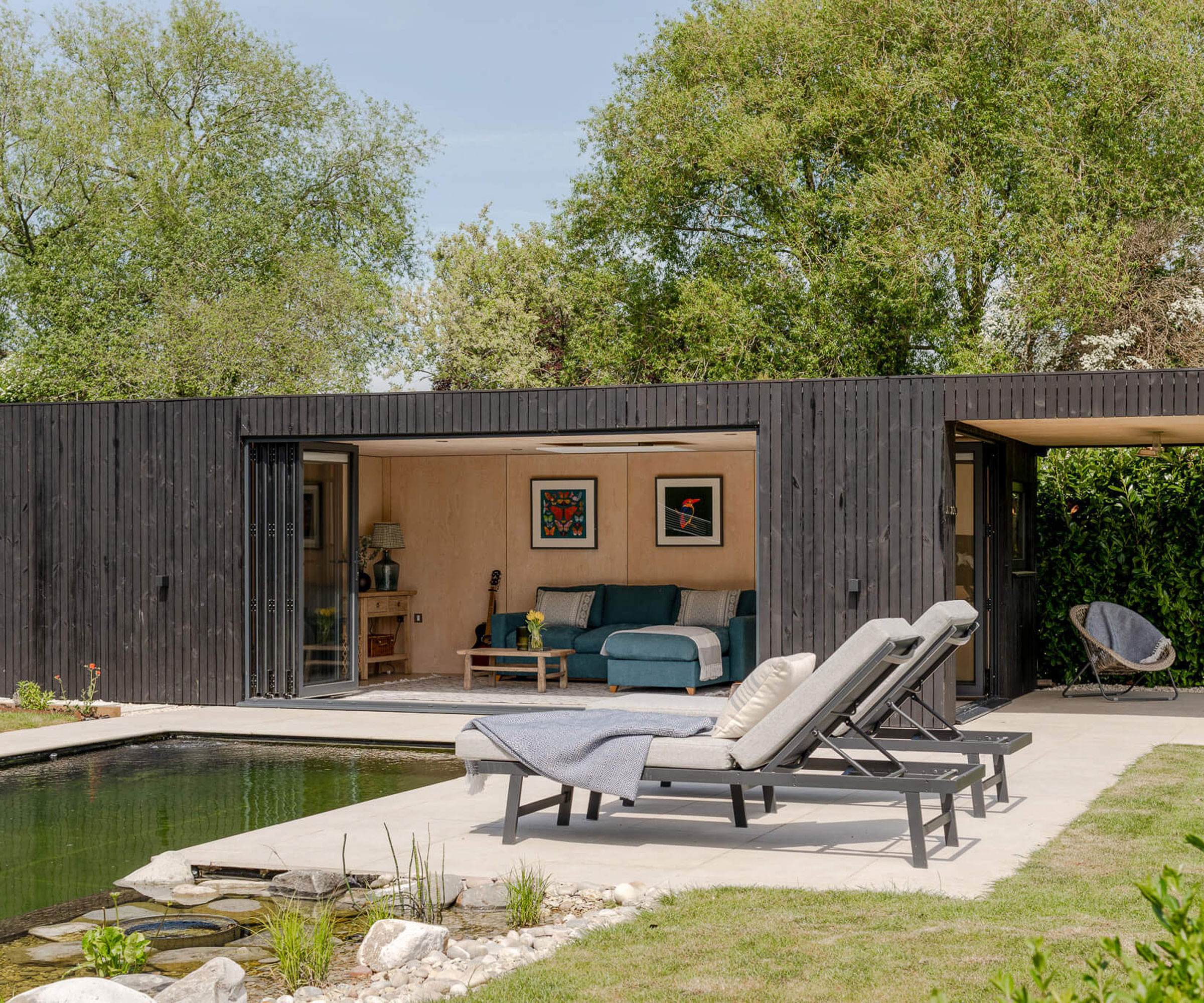 large pool house with living room area next to a natural swimming pond surrounded by trees, with patio paving, areas of planting and sun loungers