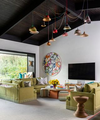 Large modern living room with dark wood paneled vaulted ceiling, white painted walls, floor to ceiling windows, seating area with green sofa and armchairs around coffee table facing a tv, artistic hanging pendant light display